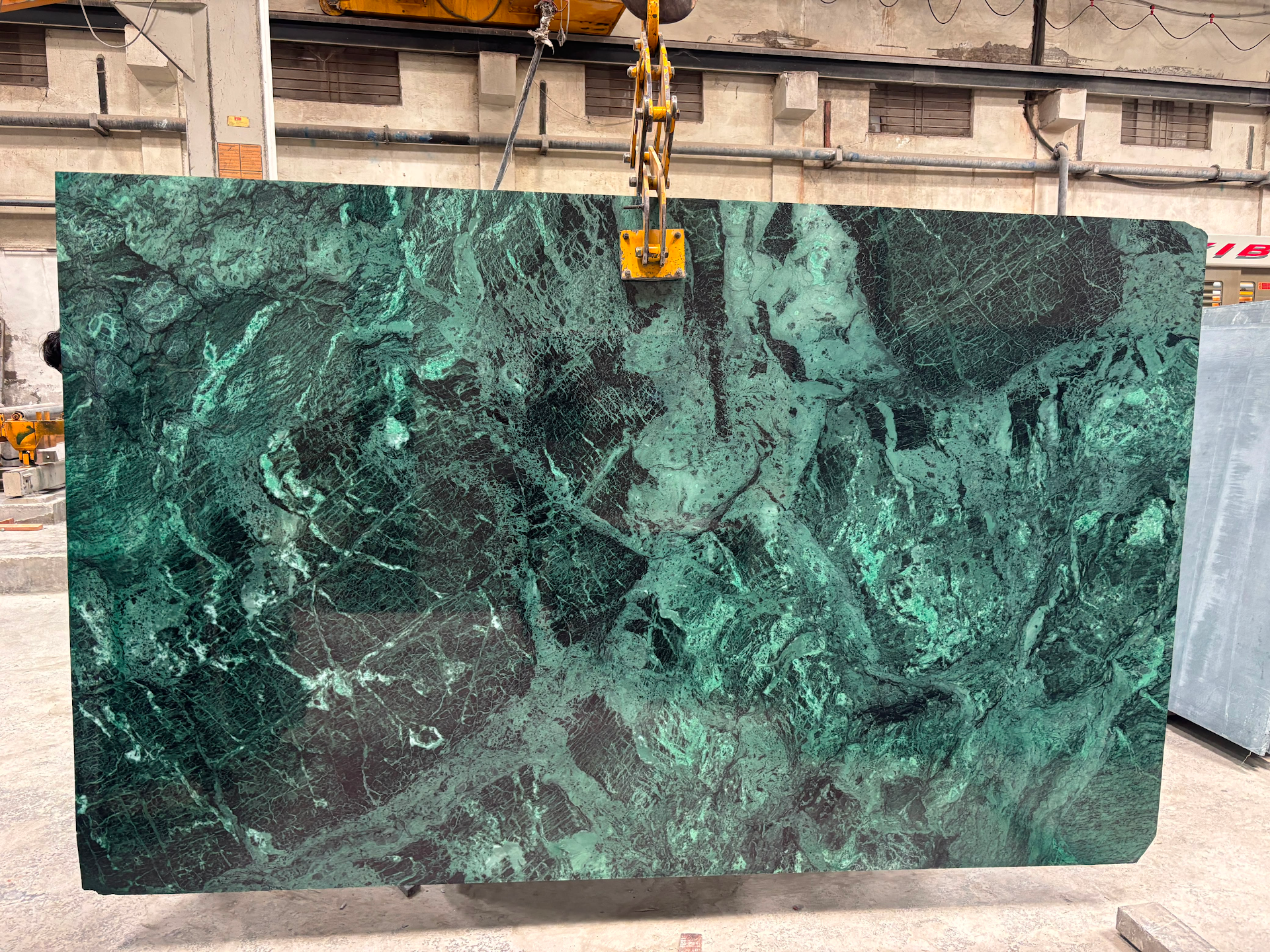 Large rectangular slab of polished green marble with complex veining, suspended by a yellow industrial crane clamp